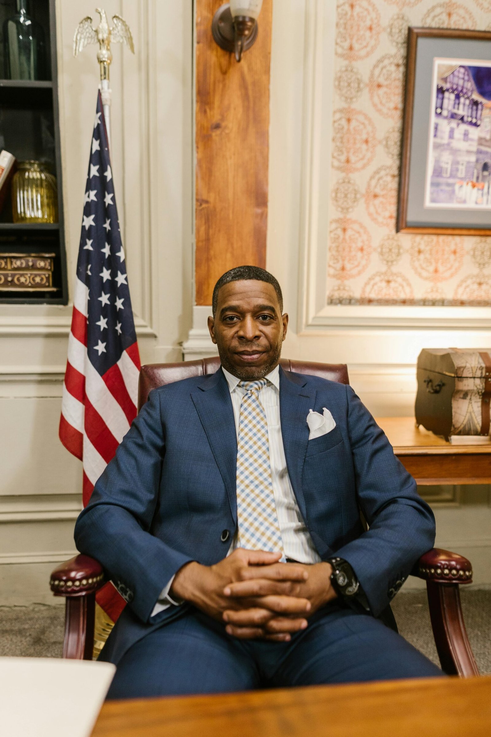 Confident man in blue suit smiling with hands together indoors. American flag in background.